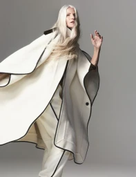 Model with long white hair wearing a cream cape with dark piping against a neutral grey studio backdrop.