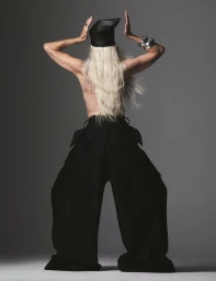 Model with long white hair wearing black wide-leg trousers and a leather cap seen from behind in a studio