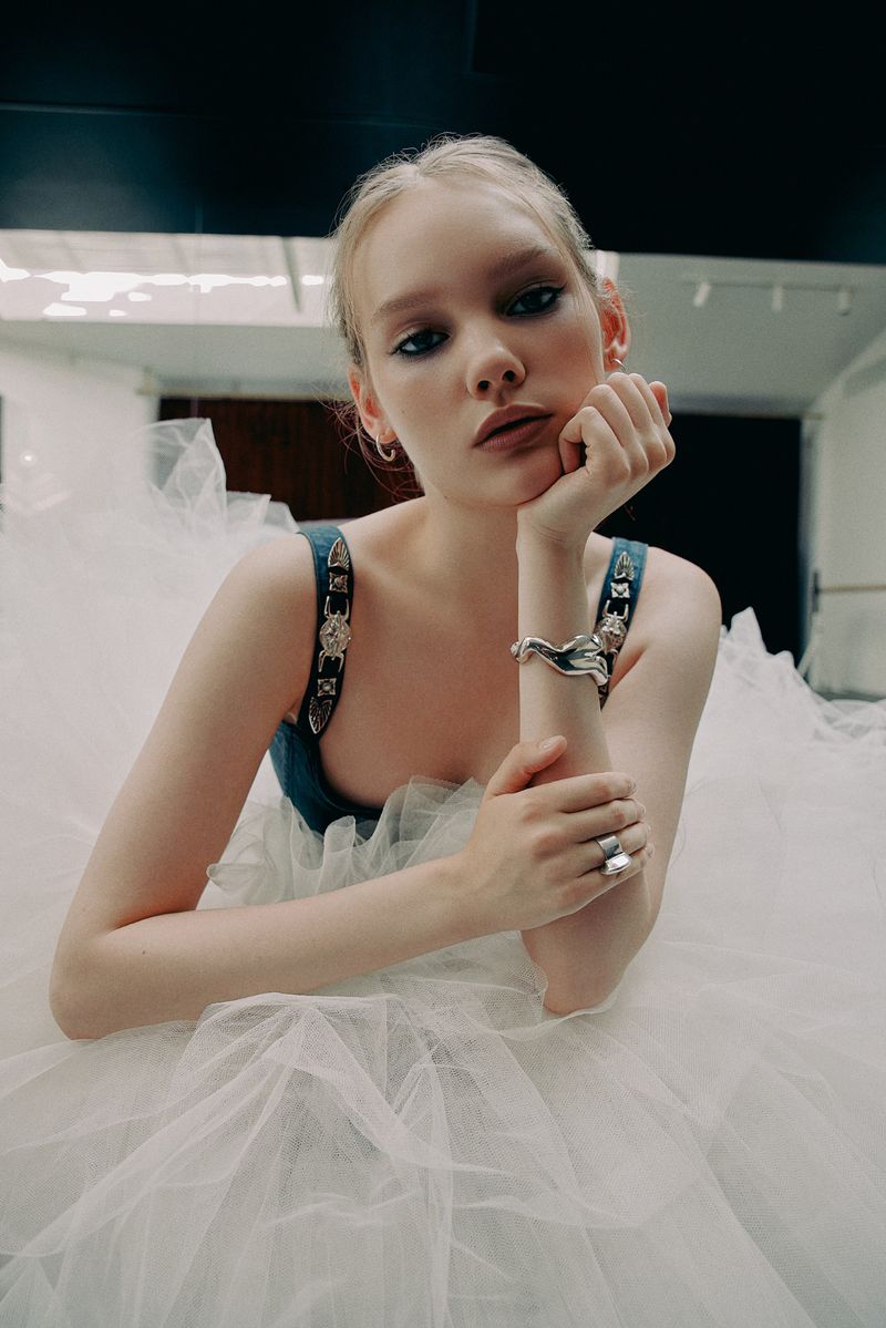 Young woman with blonde hair and blue eyes poses while sitting in a large, layered white tulle skirt, silver jewelry, and soft studio lighting.