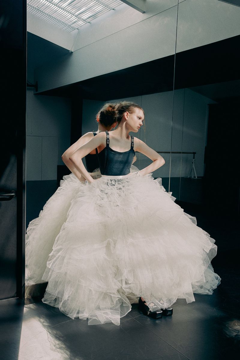 Woman in a large, frilly, white tutu stands with hands on her hips, posing next to a mirror in a dimly lit dance studio.