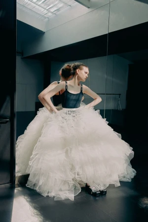 Woman in a large, frilly, white tutu stands with hands on her hips, posing next to a mirror in a dimly lit dance studio.