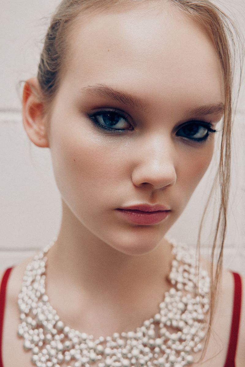 Close-up portrait of a young woman with dark eye makeup and a pearl beaded necklace against a white paneled background.