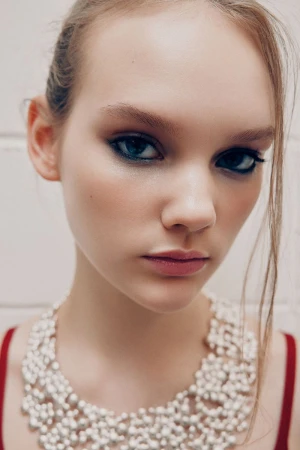 Close-up portrait of a young woman with dark eye makeup and a pearl beaded necklace against a white paneled background.
