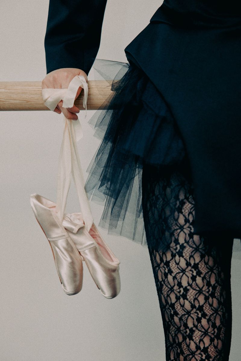 Ballet pointe shoes hang from a barre with a ballerina in a black dress and lace tights in the background, against a plain wall.