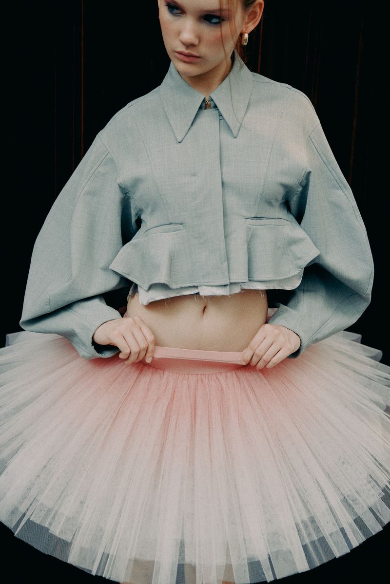 A slender young woman wearing a pale blue crop top and tutu poses against a dark background, showcasing a fashion-forward ballet-inspired look.