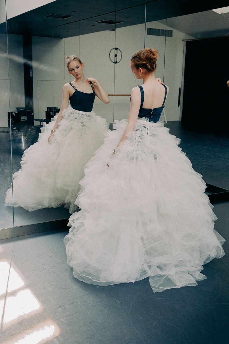 Woman in white tulle gown poses in dance studio. Mirror reflects her profile in a fashion editorial shot.