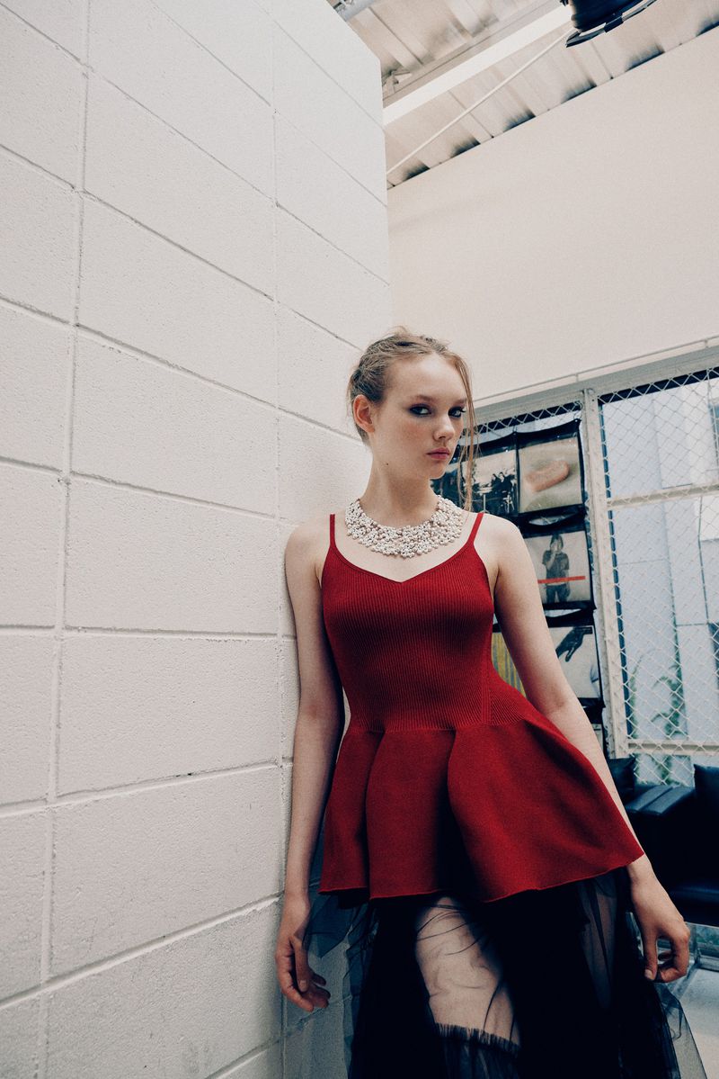 A fair-skinned woman with a red top and black skirt poses against a white brick wall near a window in a bright indoor setting.