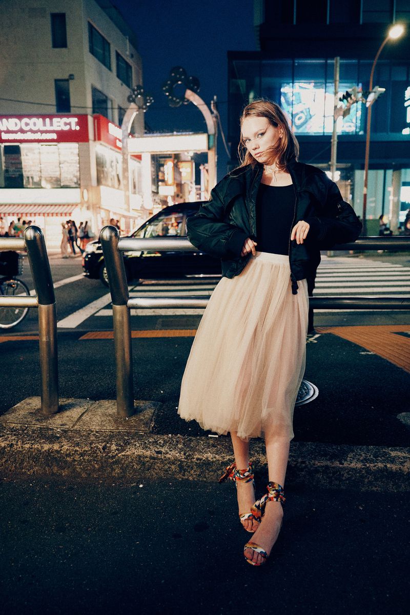 Woman in black jacket and pink tutu skirt stands on a city street at night with buildings, streetlights, and pedestrians in the background.