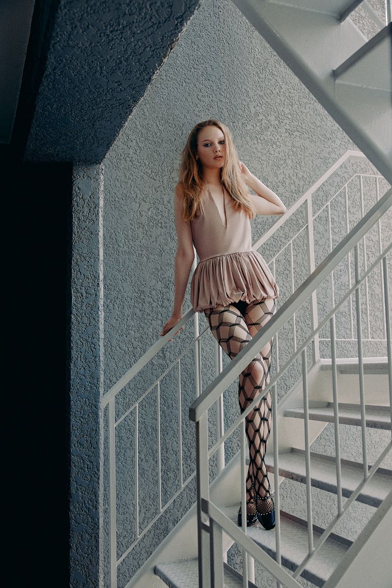 A young woman poses on a staircase wearing a beige dress, black patterned stockings, and black shoes in an indoor setting with textured walls.