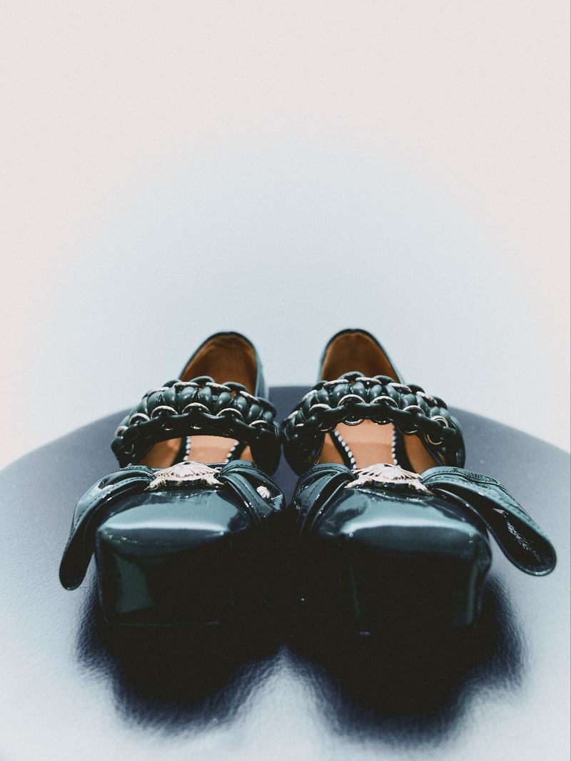 Eye-level shot of a pair of glossy black Mary Jane shoes with decorative chain trim and side bows, set against a gradient background.