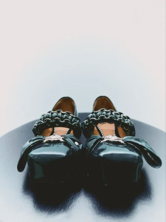 Eye-level shot of a pair of glossy black Mary Jane shoes with decorative chain trim and side bows, set against a gradient background.