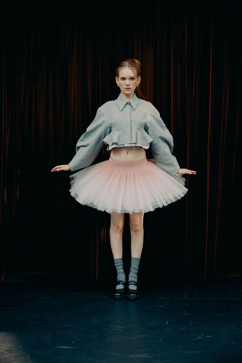Young woman in a tutu and jacket stands in a dance studio, against a dark curtain backdrop. The studio setting enhances the dancer's poised stance.