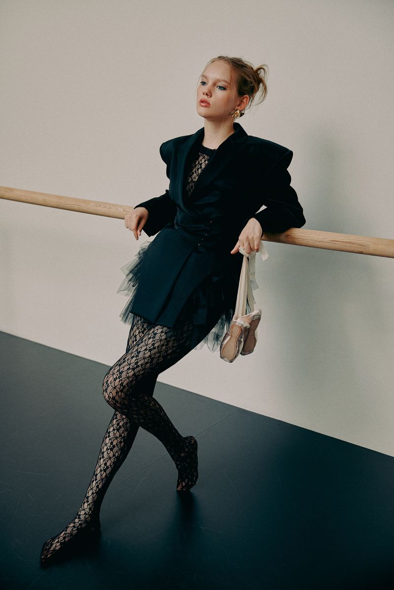 Young woman in black jacket and lace tights holds ballet shoes, leaning against barre in a photo studio setting. Fashion editorial.