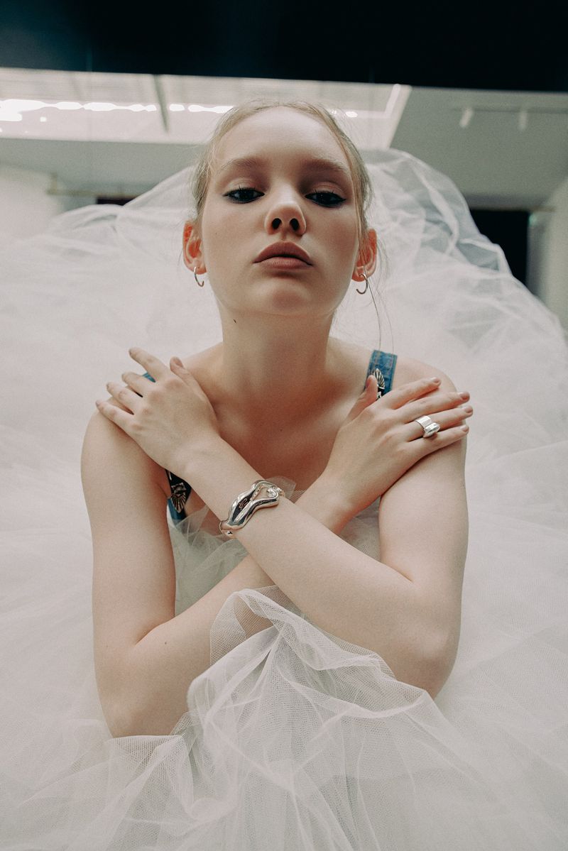 Portrait of a young woman in a white dress with a tulle skirt, arms crossed, wearing silver jewelry, in an indoor setting.