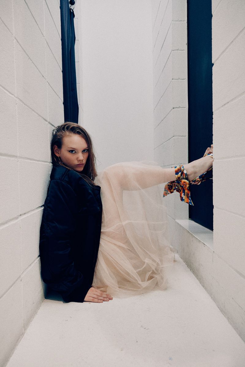 Woman in a sheer dress and black jacket poses in a white tiled space, creating a striking contrast and a modern, artistic aesthetic.
