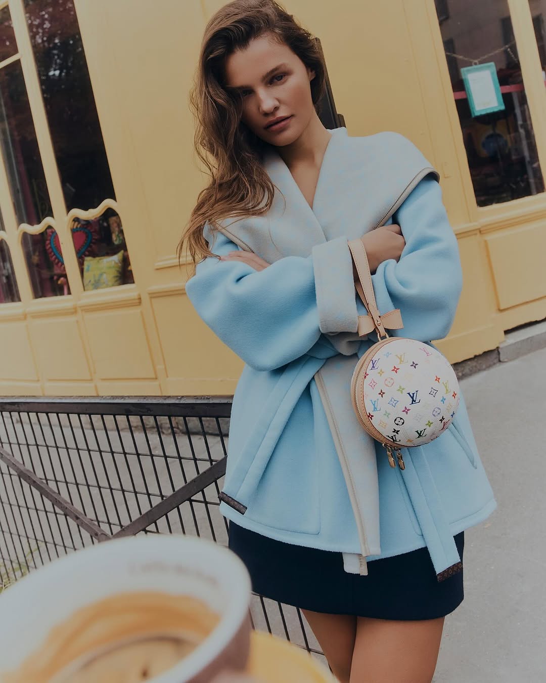 A stylish woman in a blue coat and black skirt poses outdoors with a Louis Vuitton handbag in front of a yellow building.