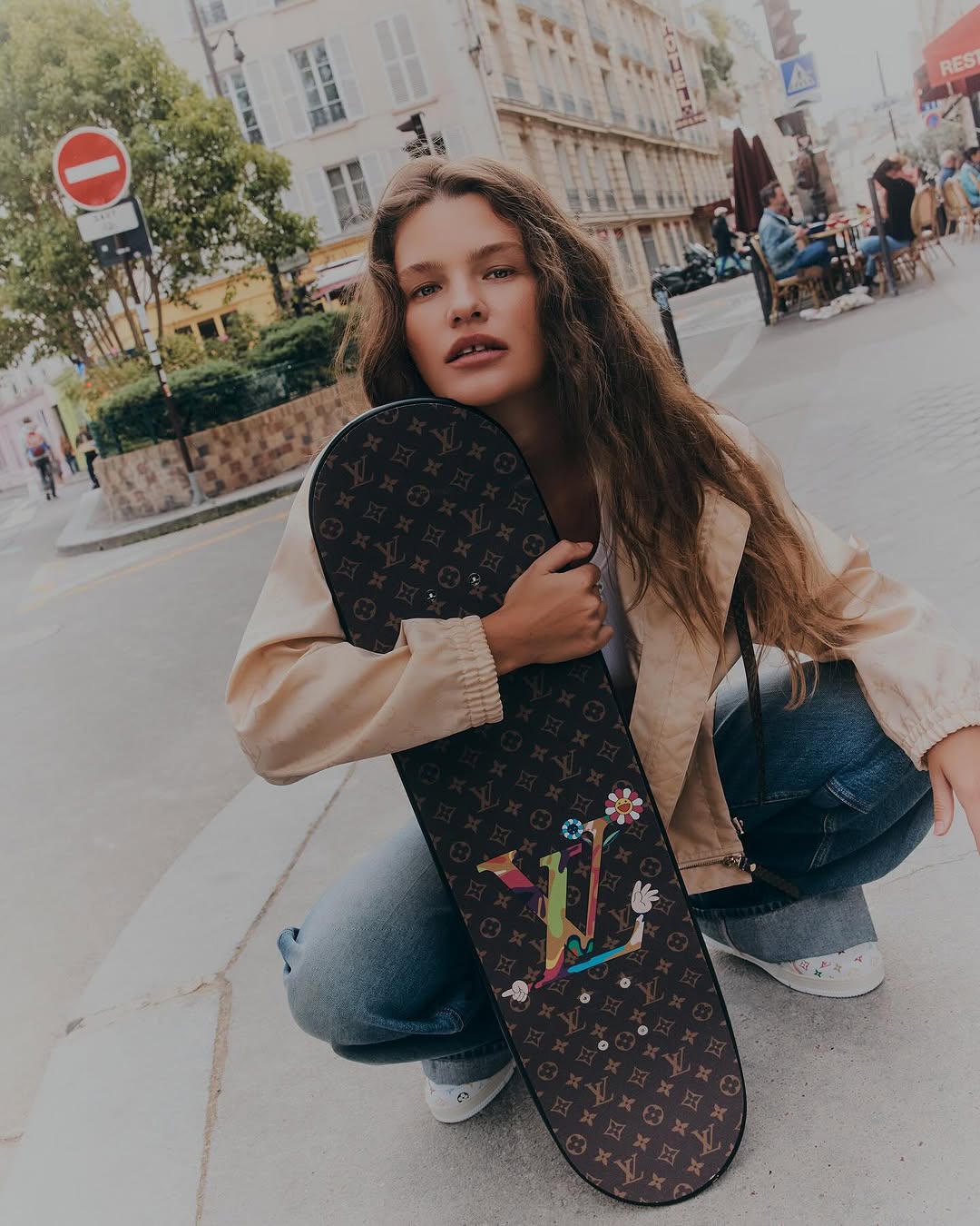 A woman crouches holding a Louis Vuitton skateboard, pictured on a city sidewalk with buildings and a no entry sign in the background.