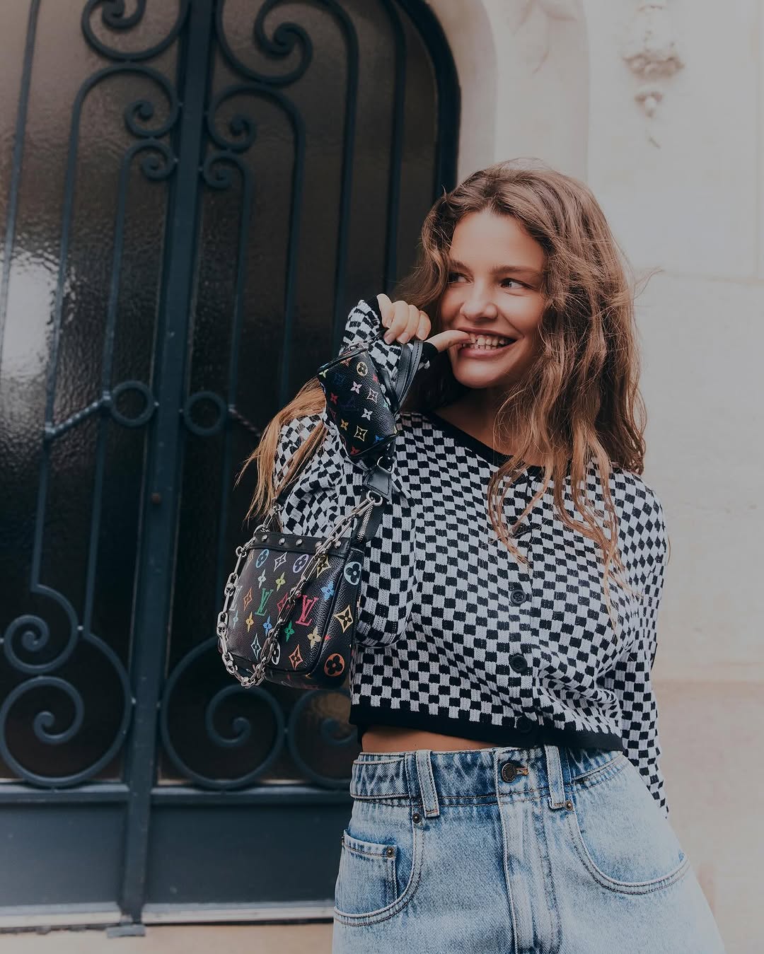 A stylish young woman in a checkerboard cardigan and light jeans, posing with a patterned bag in an outdoor, urban setting.