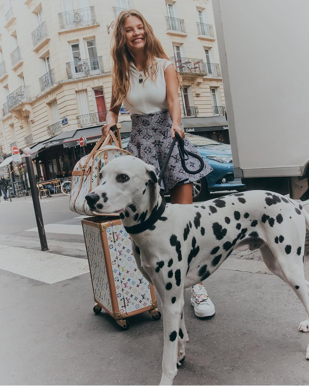 Woman walking a dalmatian dog in a European city, with luggage and buildings in the background, capturing a moment of urban lifestyle.