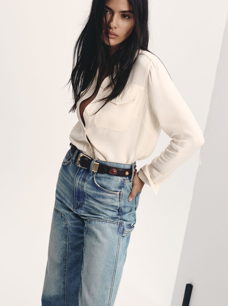A woman poses in a white button-down shirt and light wash jeans with a decorative belt against a neutral backdrop.