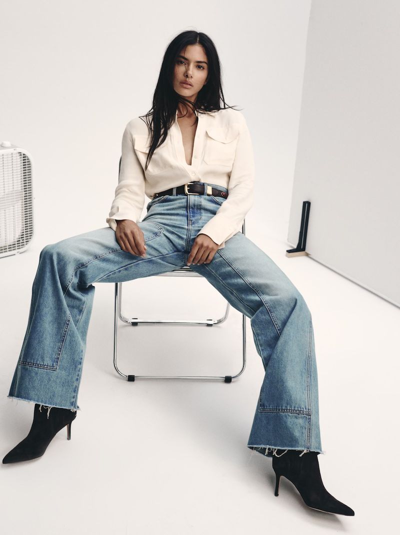 Fashionable woman in light shirt and wide-leg jeans sits on metal chair in bright studio. Dark hair, black boots. Fan and panel backdrop.