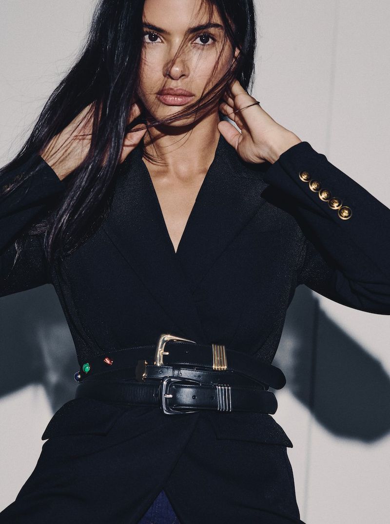 A woman poses indoors in a black blazer cinched with belts; a fashion portrait featuring dark hair and fair skin against a plain background.