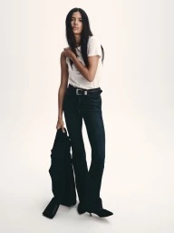 Model wearing a white t-shirt and dark denim flare jeans holding a black jacket against a plain light background