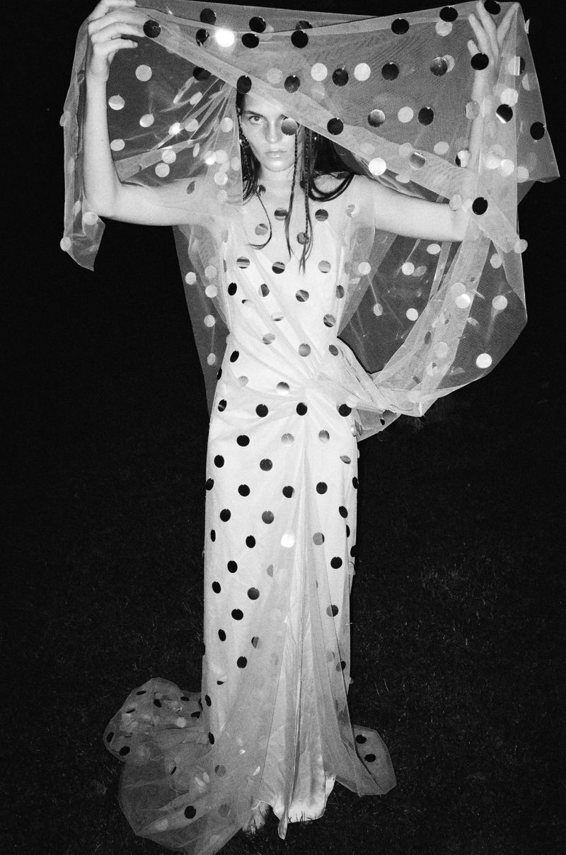 Woman in dotted dress and veil poses in black and white. Fashion shot with sheer fabrics and polka dot pattern in a dark setting.
