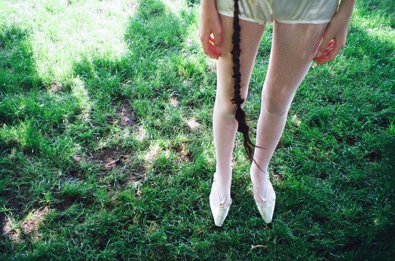 Person wearing tights and heels stands on green grass with hair braid visible. Outdoor setting with natural light in a close-up shot.