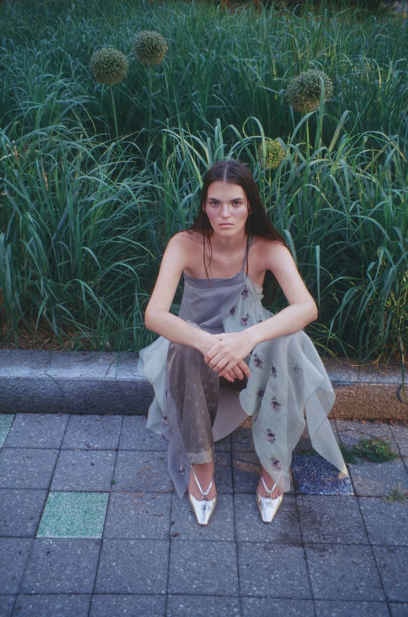 A young woman with long brown hair and silver shoes sitting on a stone ground with long green grass and flowers in the background.