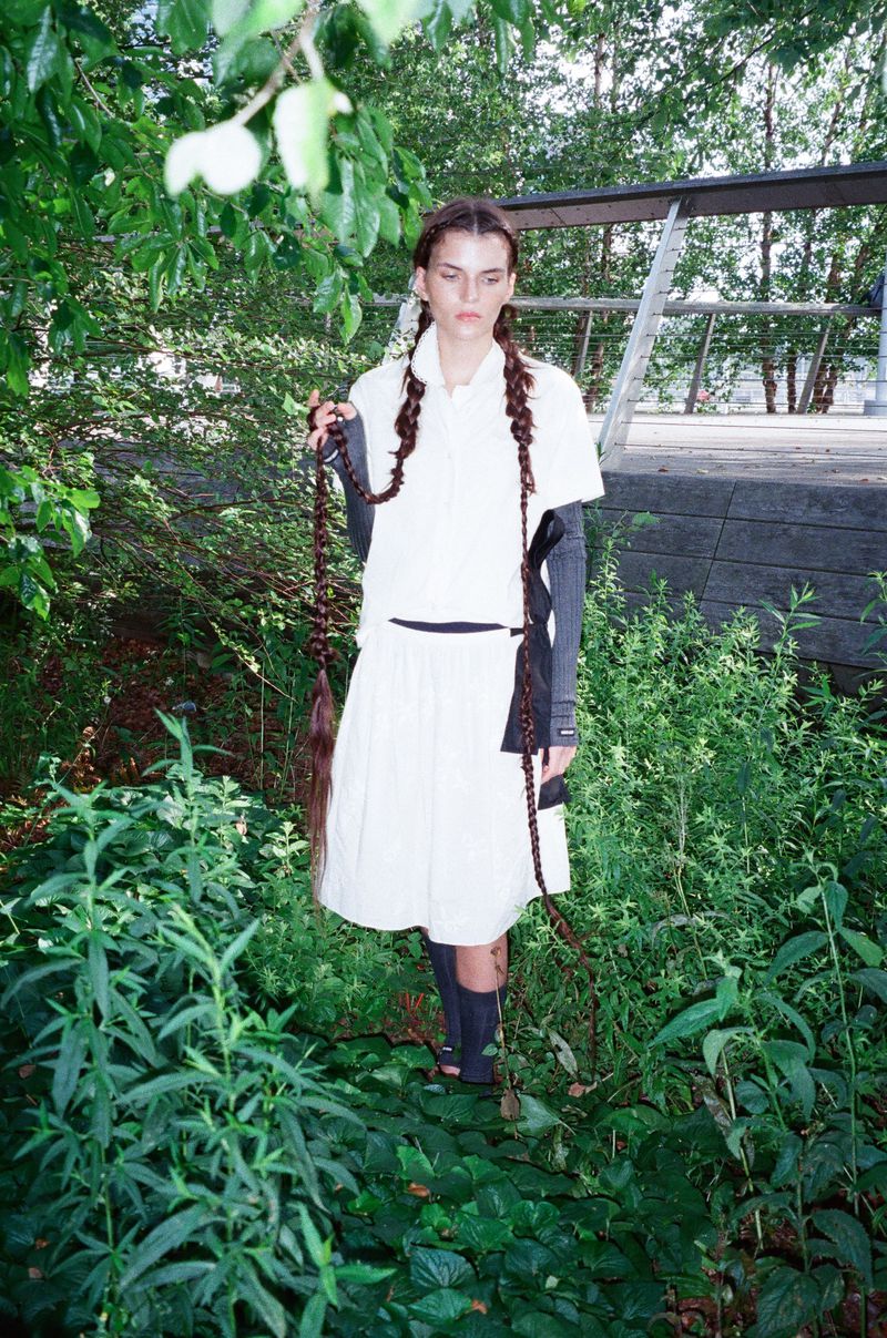 A young adult woman in a white dress and long braids stands in a park setting surrounded by lush green foliage and trees.