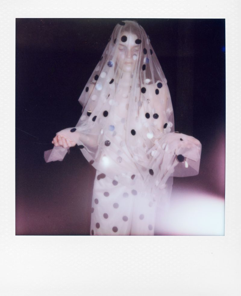 Woman in a polka-dot dress and veil stands in a dark space with light flares. Fashion shot with a unique retro Polaroid aesthetic.