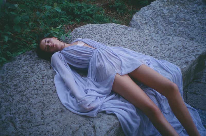 A young woman in a lilac dress lies on a rock, surrounded by greenery, in a visually striking and ethereal outdoor fashion shot.