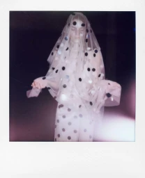 Model in a polka dot sheer veil and dress standing against a dark background in a polaroid style photograph