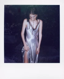 Polaroid photograph of a woman wearing a glossy silver silk slip dress standing outdoors at night amidst dark foliage.