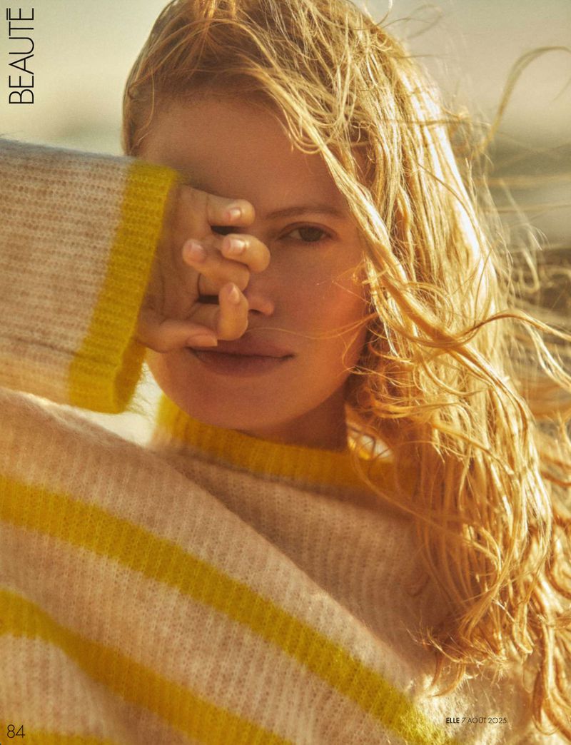 Close-up portrait of a woman with long blonde hair wearing a striped yellow and white sweater, covering her eyes with her hand.