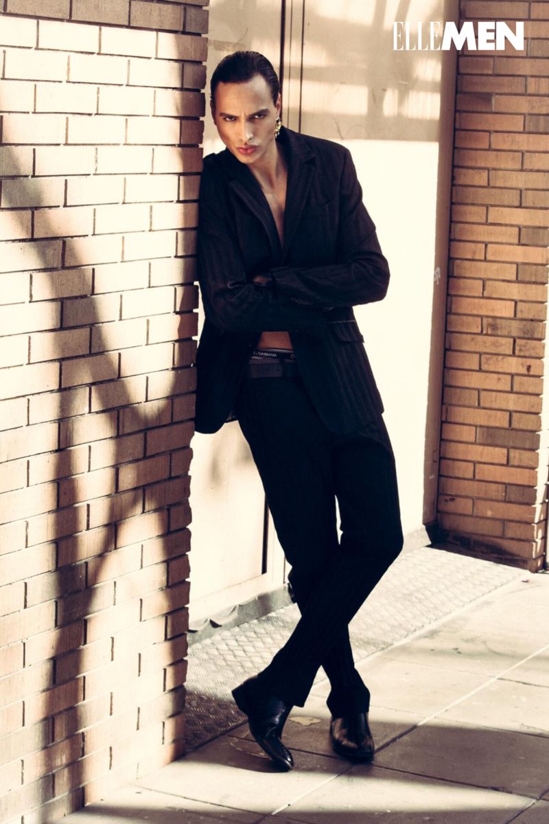 A male model poses in a black suit against a brick wall. The lighting creates dramatic shadows, highlighting the fashion editorial style.
