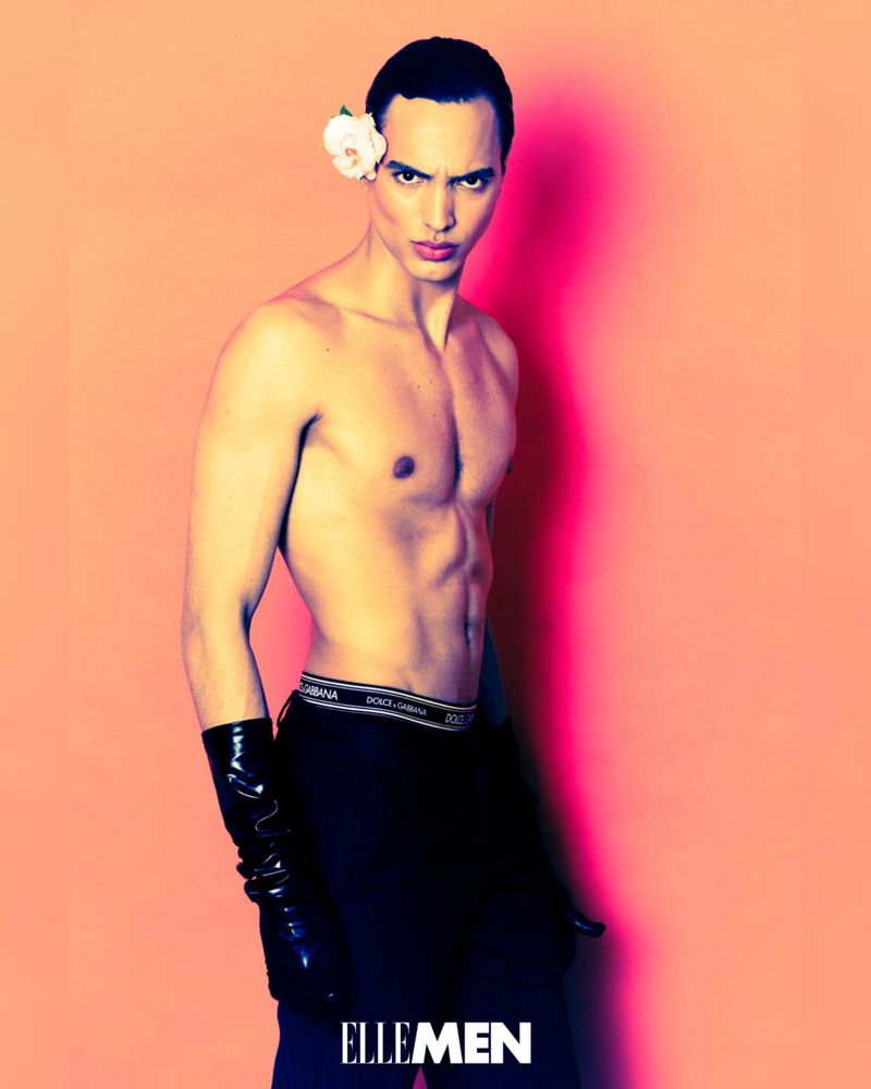 Shirtless man with rose in hair poses in front of pink background wearing black bottoms and long gloves for Elle Men editorial.