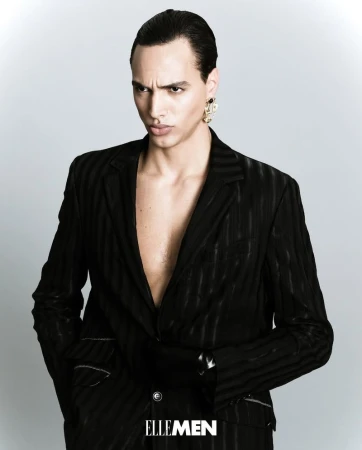 Man in black suit with open blazer poses in a fashion editorial shot against a neutral grey backdrop wearing gold statement earrings.