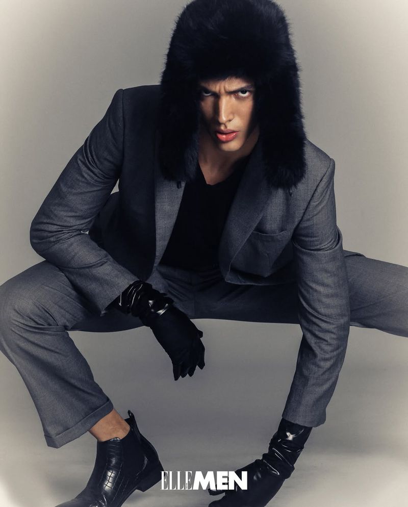Man poses in gray suit and black fur hat for an Elle Men fashion editorial against a gray background in a photo studio.
