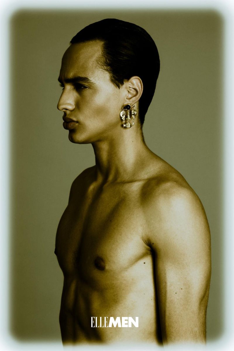 Close-up studio shot of a shirtless adult male model in profile with dark hair and olive skin, wearing large ornate gold earrings.