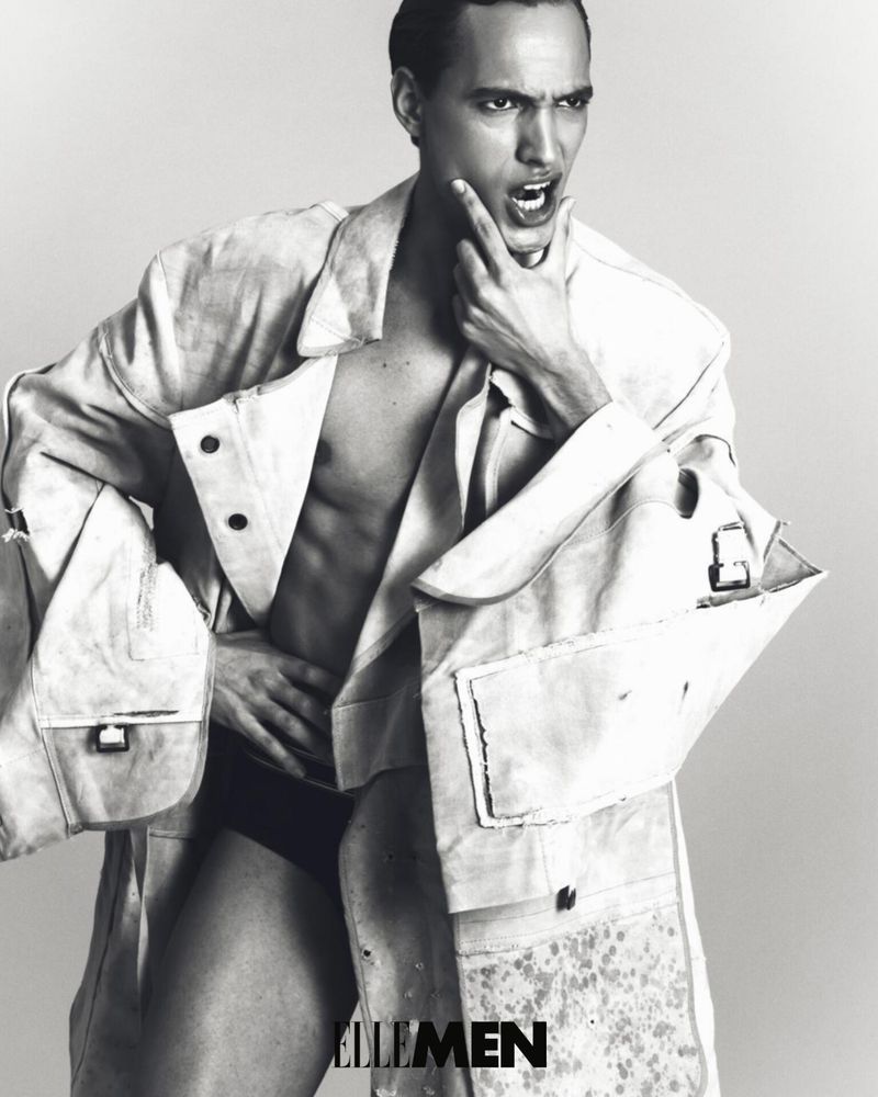 Black and white studio shot of a male model with an open coat revealing his chest, posing with hand on his chin.