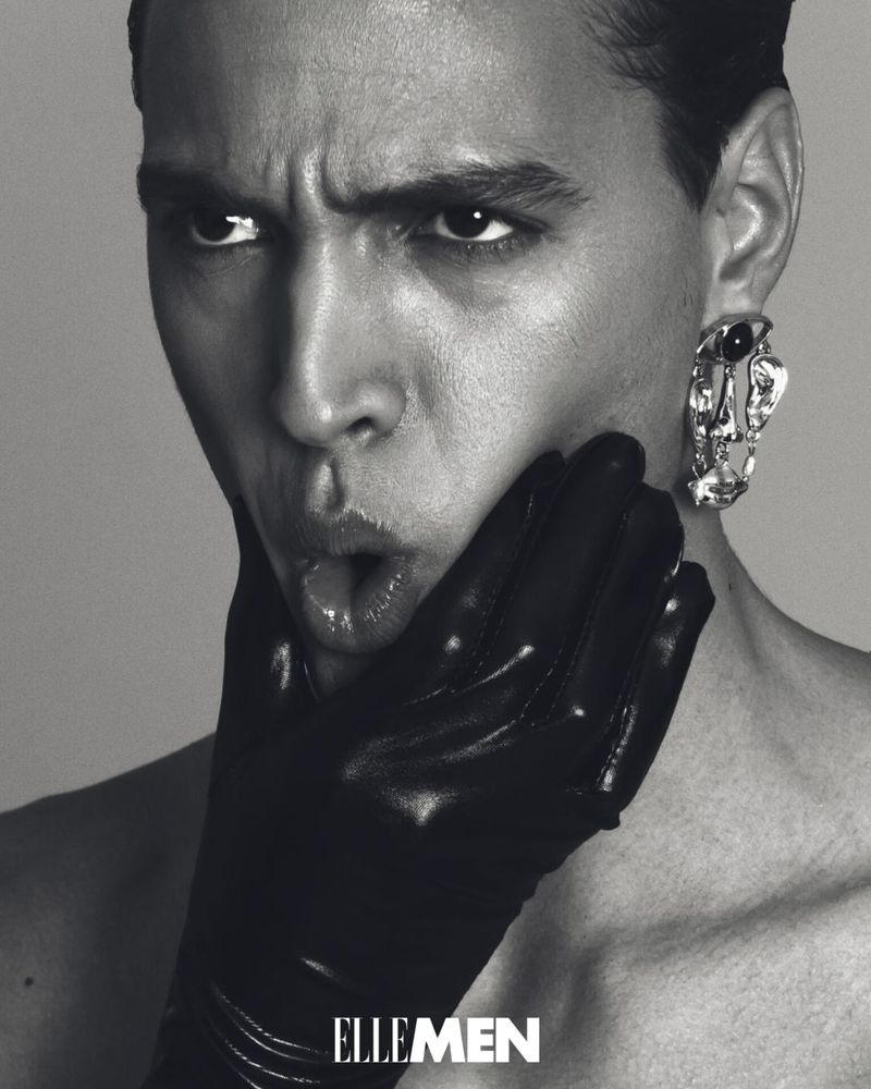 Monochromatic portrait of a man wearing a black glove and earring, making a playful expression in front of a solid light-grey background.