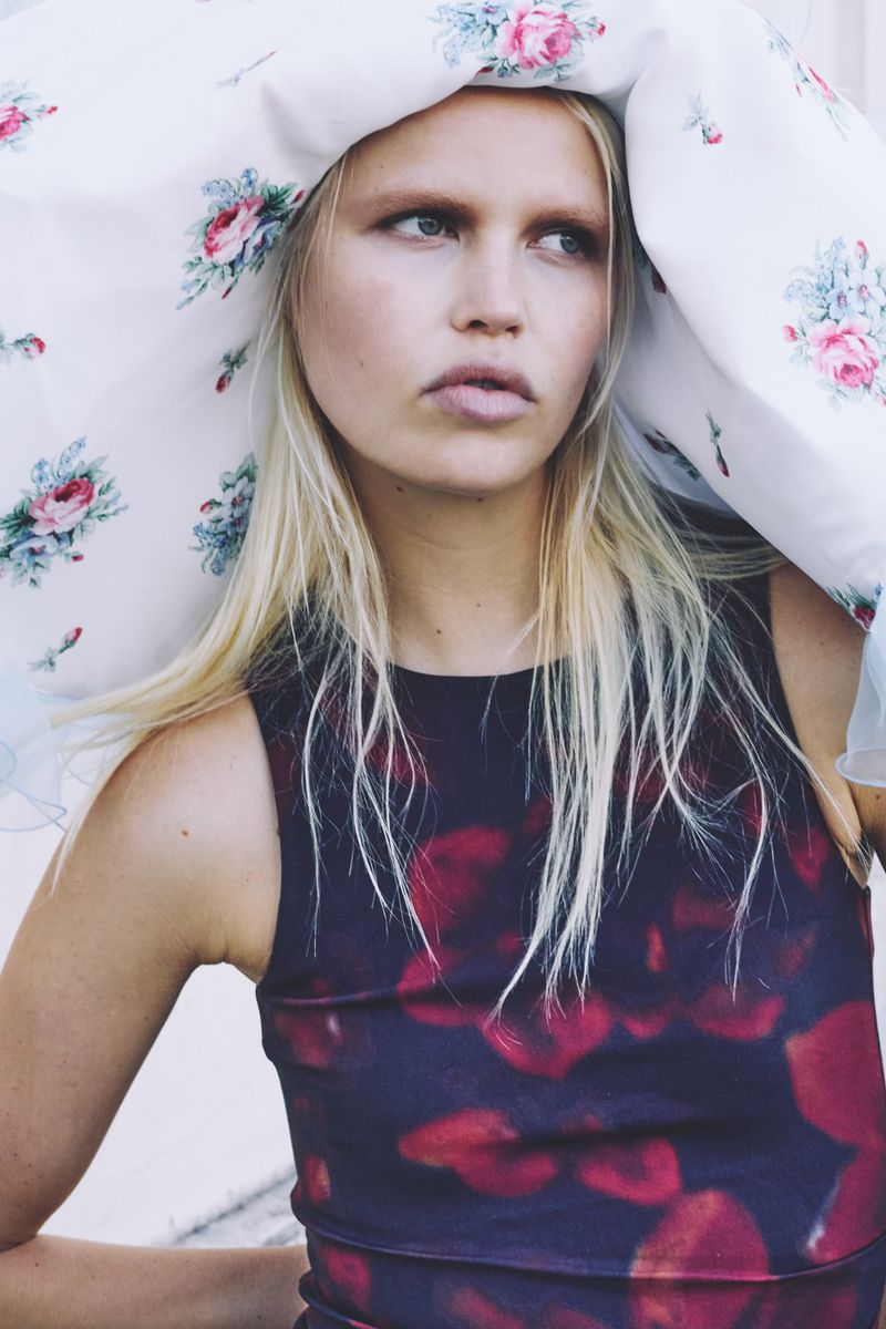 Close-up of a fair-skinned, blonde woman wearing a dark dress with red patterns, a floral-patterned cloth above her head, looking off-camera.