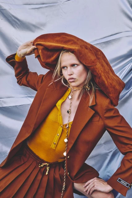 Woman in brown blazer and skirt with yellow top, accessorized with a chain necklace and earrings, posing against a silver backdrop.