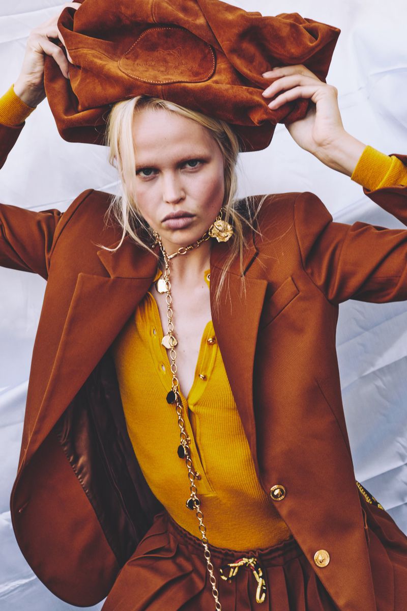 A fair-skinned woman poses in a studio wearing a brown blazer, a yellow ribbed top, and holding a brown suede jacket overhead.