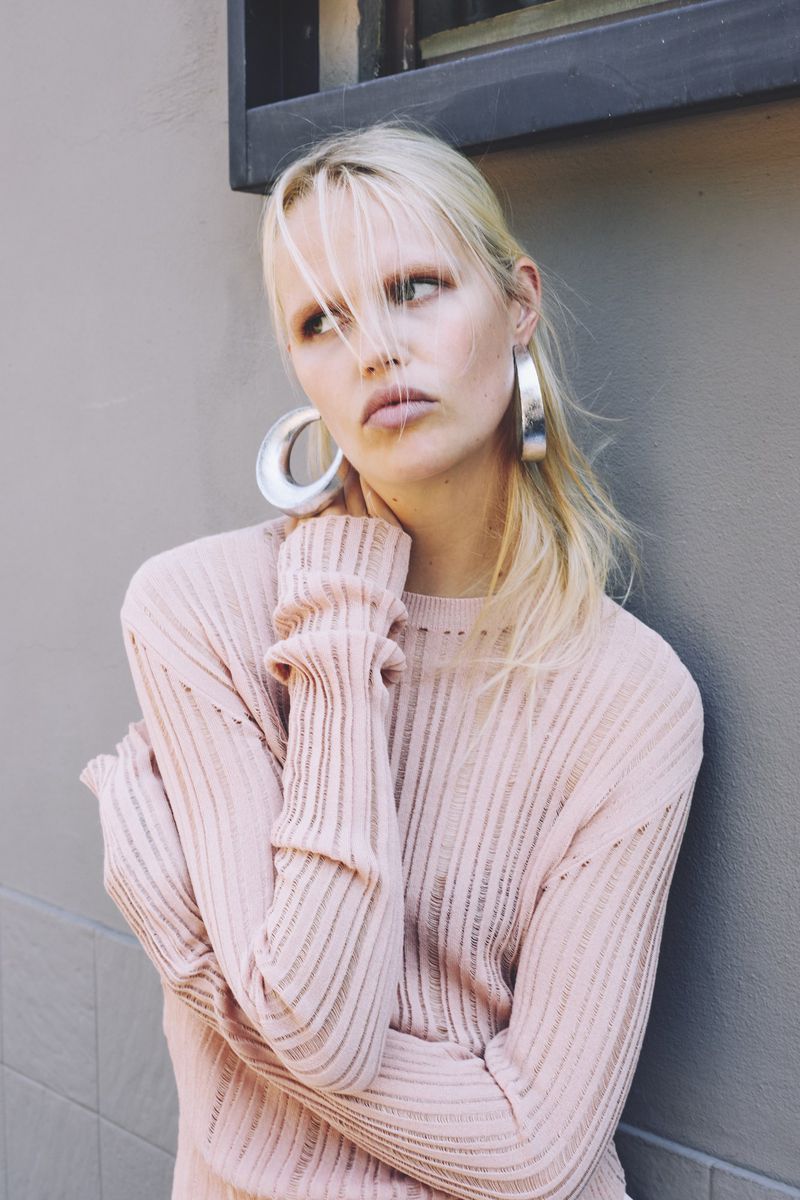 Portrait of a young adult woman in a light pink ribbed sweater and silver hoop earrings against a neutral grey background. Fashion shot.