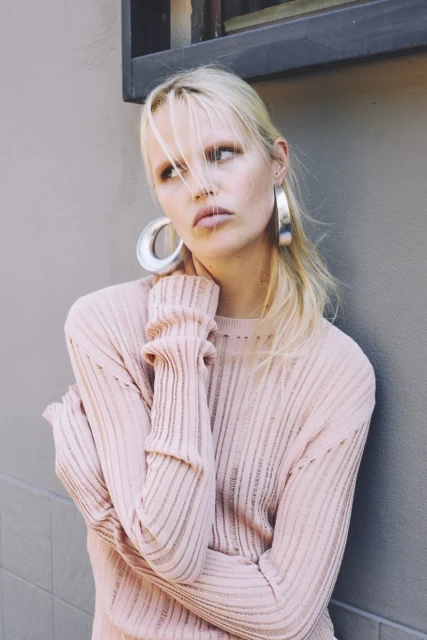 Portrait of a young adult woman in a light pink ribbed sweater and silver hoop earrings against a neutral grey background. Fashion shot.