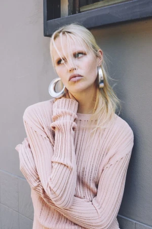 Portrait of a young adult woman in a light pink ribbed sweater and silver hoop earrings against a neutral grey background. Fashion shot.