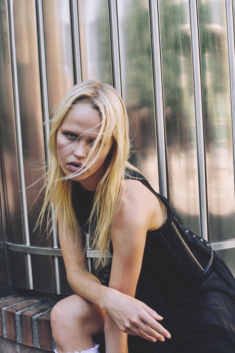 Young woman with blonde hair poses sitting against a metal wall. She wears a black dress and has a neutral facial expression.